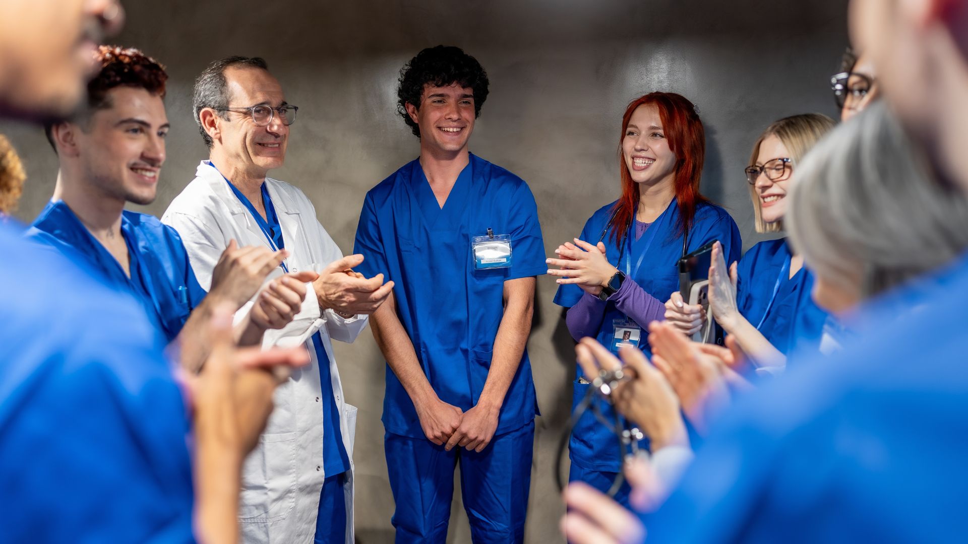 A group of healthcare workers in blue scrubs and one clinician in a white lab coat stand in a circle indoors, smiling and applauding.