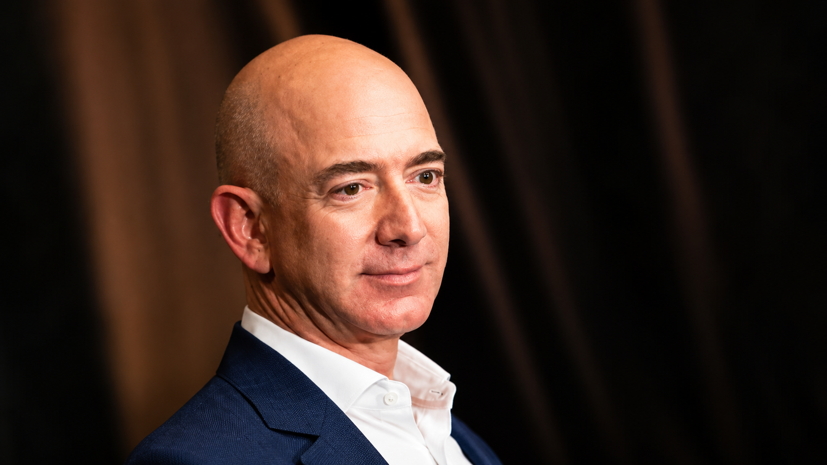 Amazon founder Jeff Bezos in a blue suit jacket and open-collar white shirt posing against a dark background.