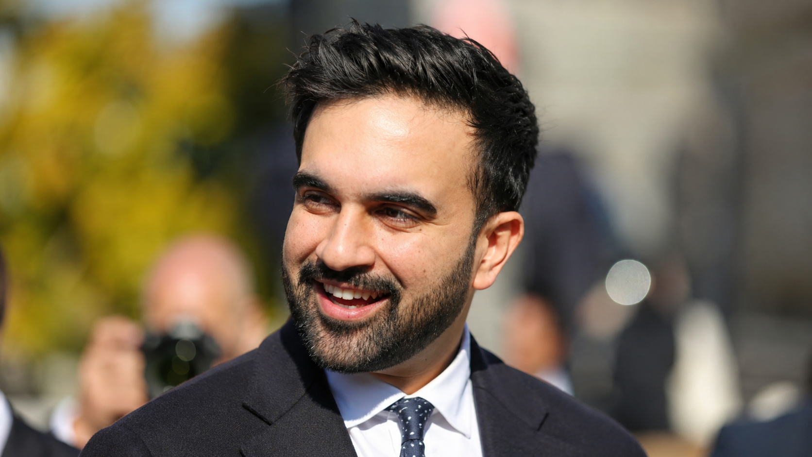 Zohran Mamdani smiling while walking outdoors.