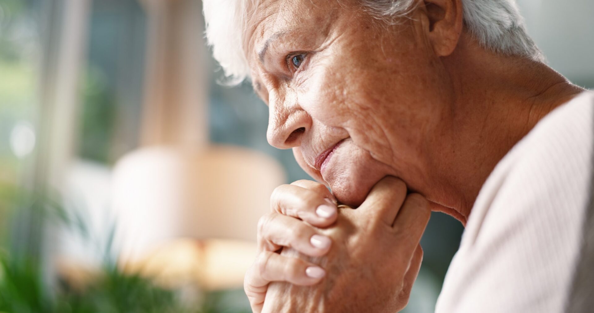 Elderly woman resting her chin on clasped hands in a thoughtful moment at home.