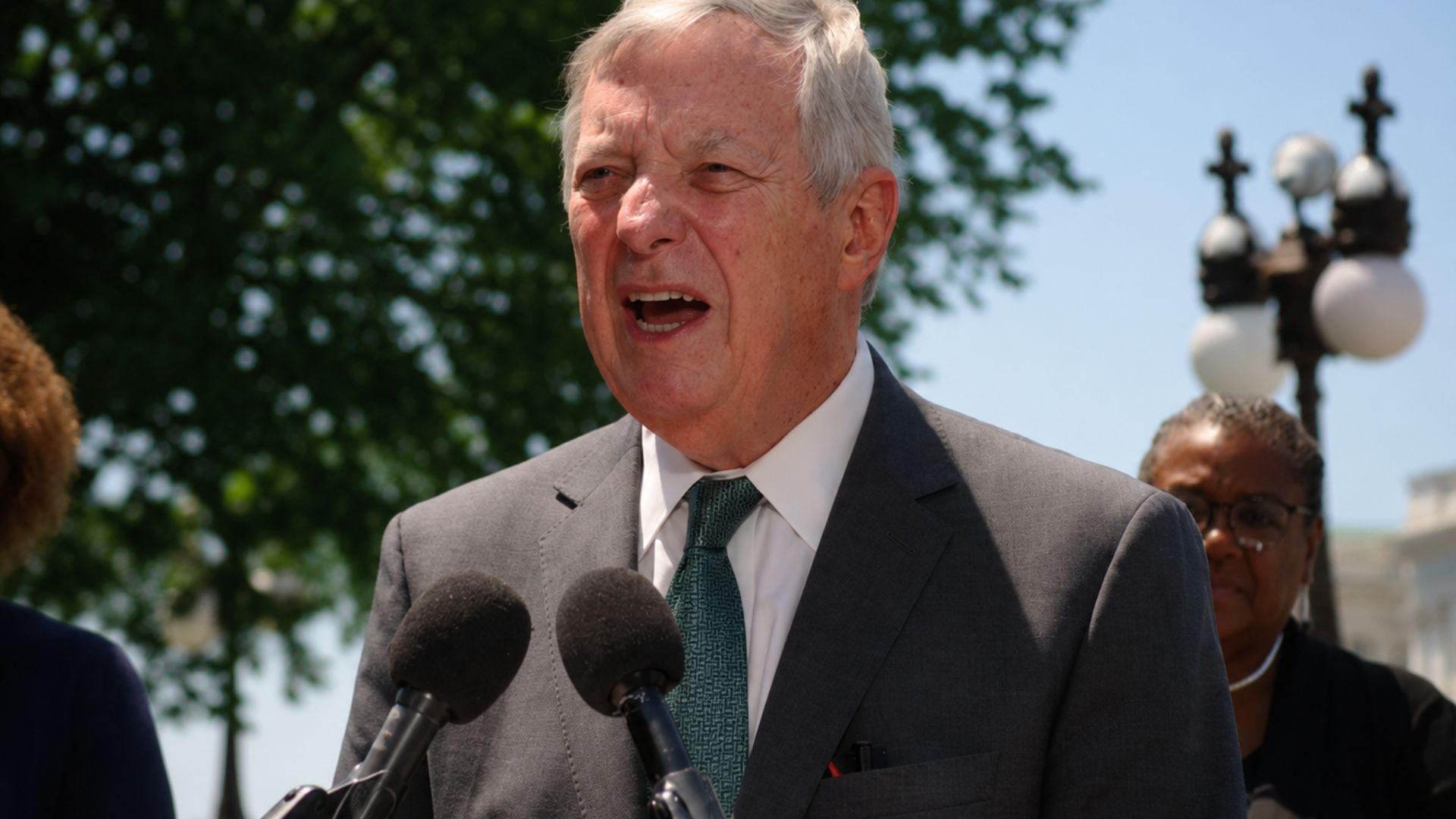 Man in a suit speaking at microphones during an outdoor news conference.