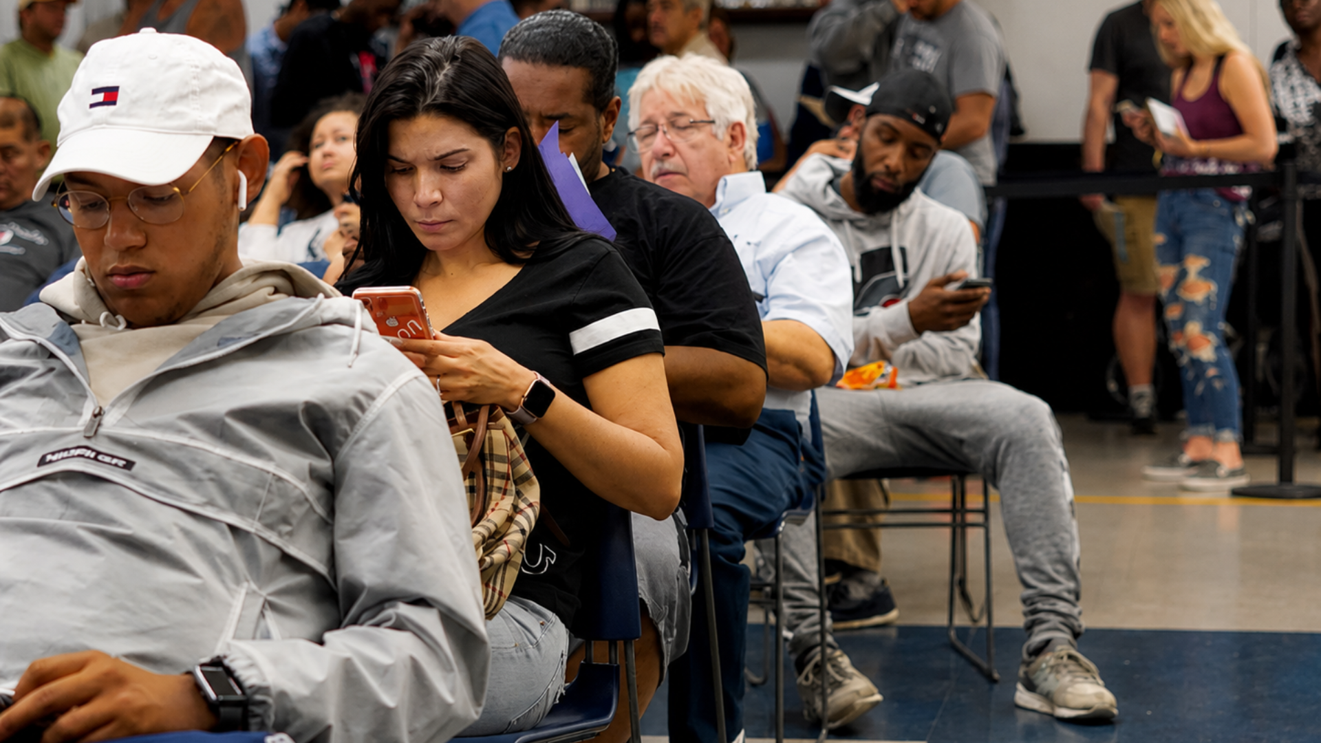 People wait in line at a motor vehicle office, many looking at their phones while seated.