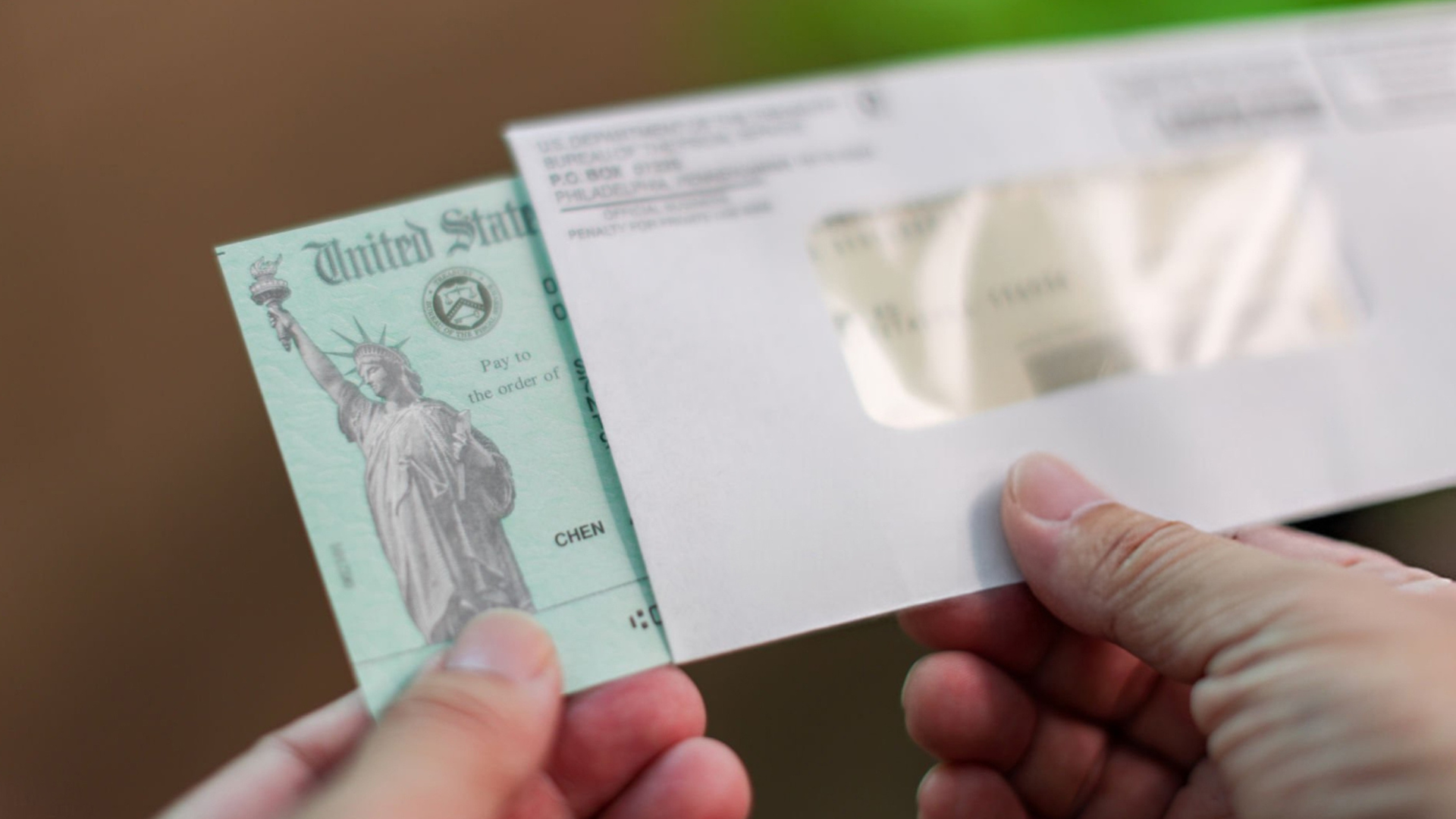 A close-up of hands pulling a green United States Treasury check out of a white windowed envelope.