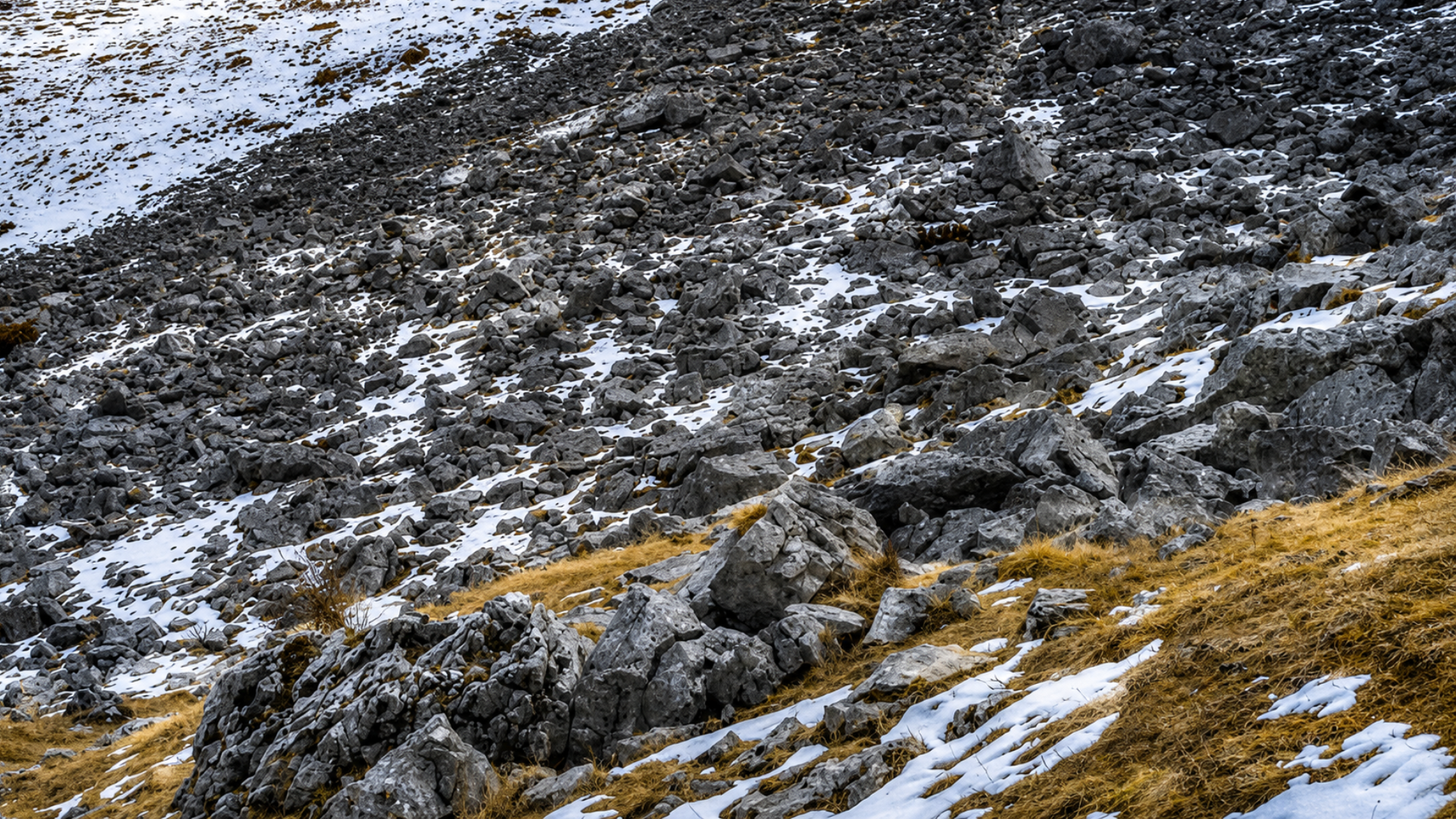 A rocky mountain slope showing light, sparse snowpack over exposed gray stones and dry vegetation.