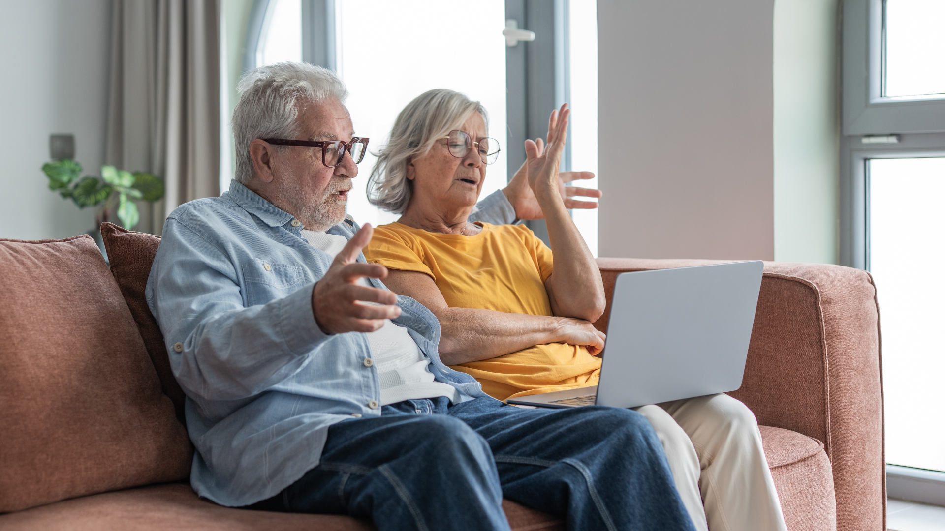 An older couple sitting on a couch, gesturing with frustration while looking at a laptop screen together.