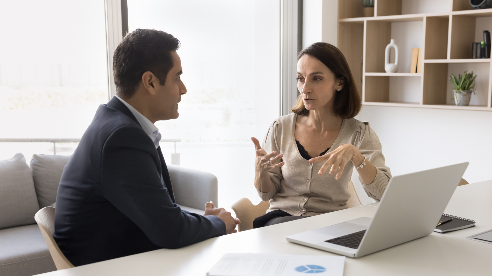 A woman gesturing with her hands while having a serious discussion with a man in a suit across a white desk.