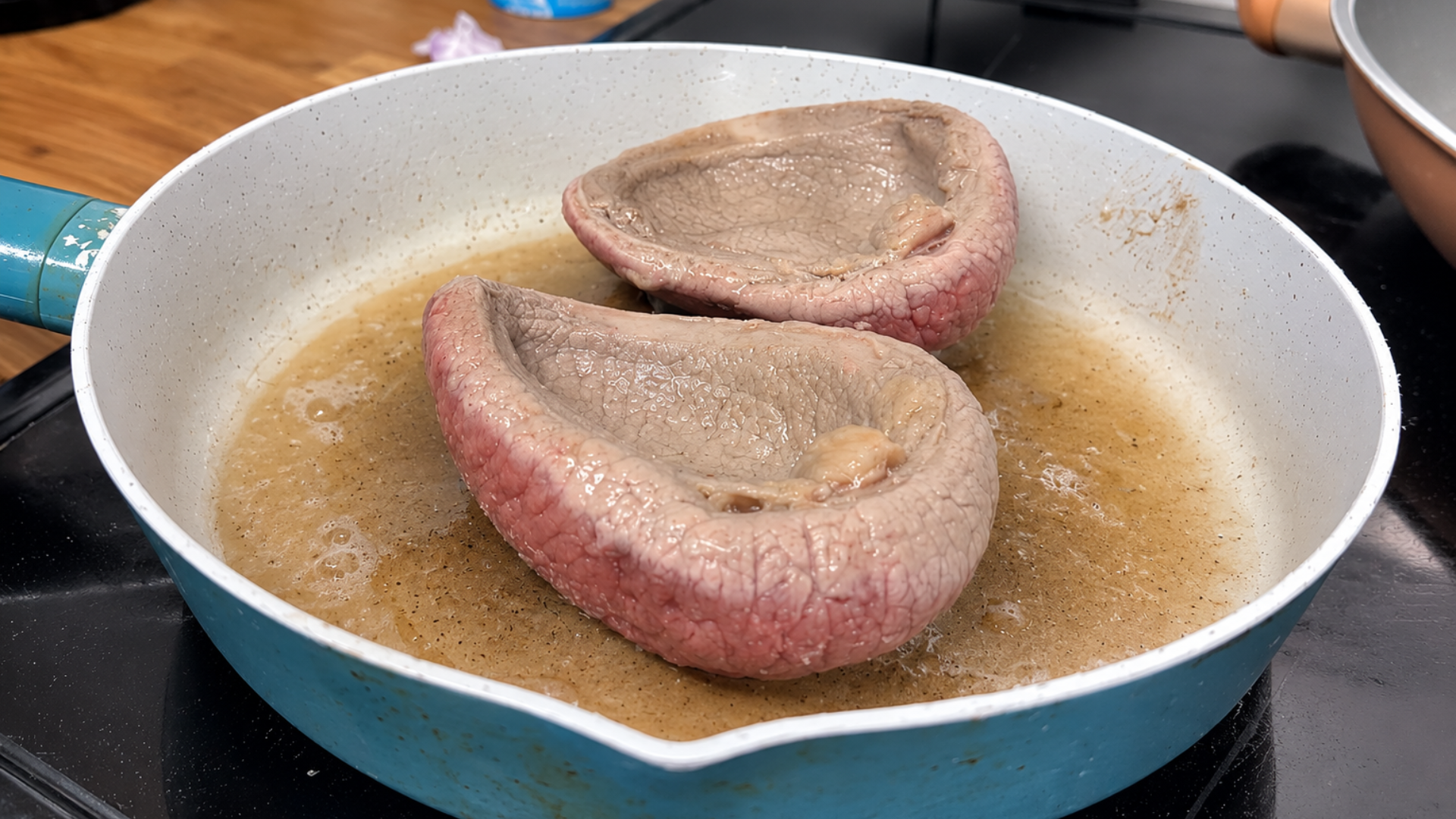 Two beef tongues simmering in butter or cooking juices in a frying pan on a stovetop.
