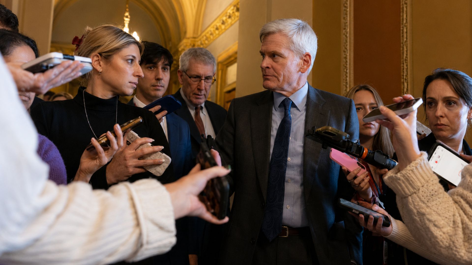 Man in a suit speaking with reporters as journalists hold microphones and smartphones during an interview.