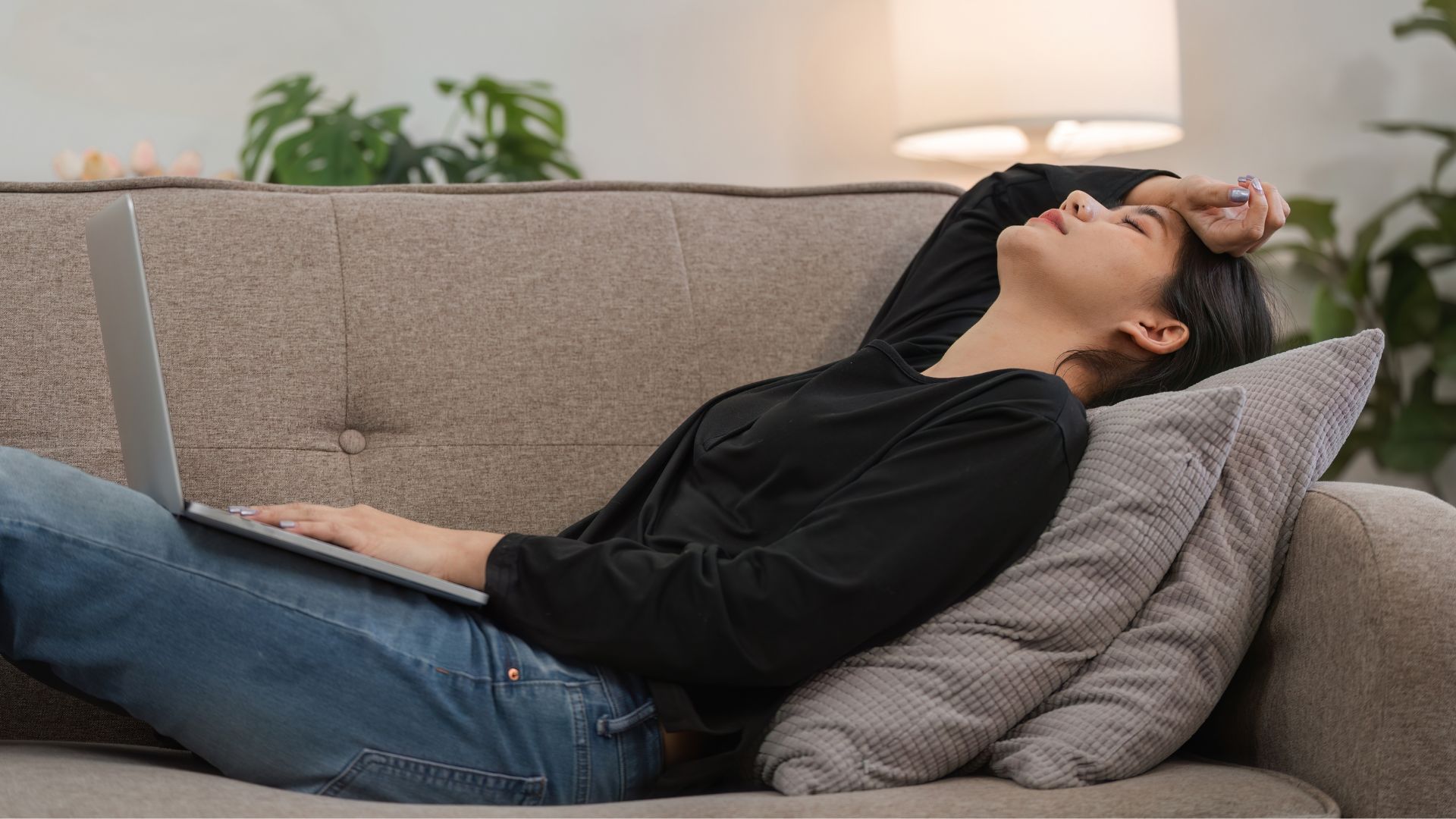 Woman lying on a couch with a laptop and resting a hand on her forehead.
