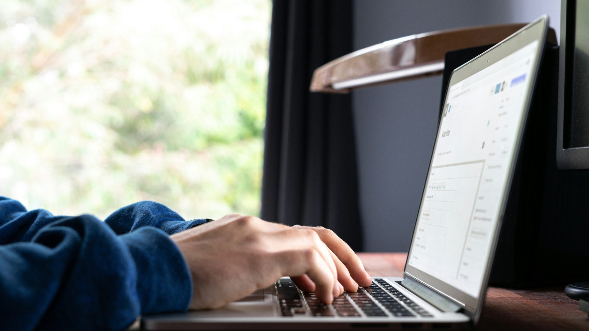A person types on a laptop displaying a webpage with an online form while seated at a desk near a window.