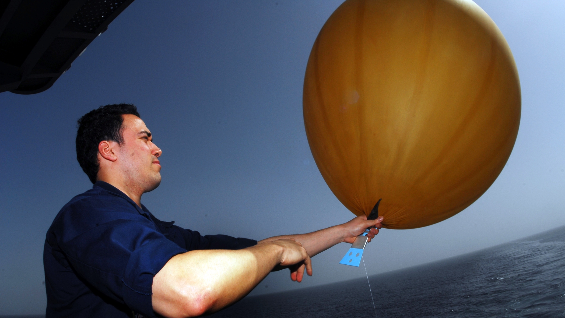 Weather specialist on the deck of a ship prepares to release a large orange weather balloon over the ocean under a cloudy sky.