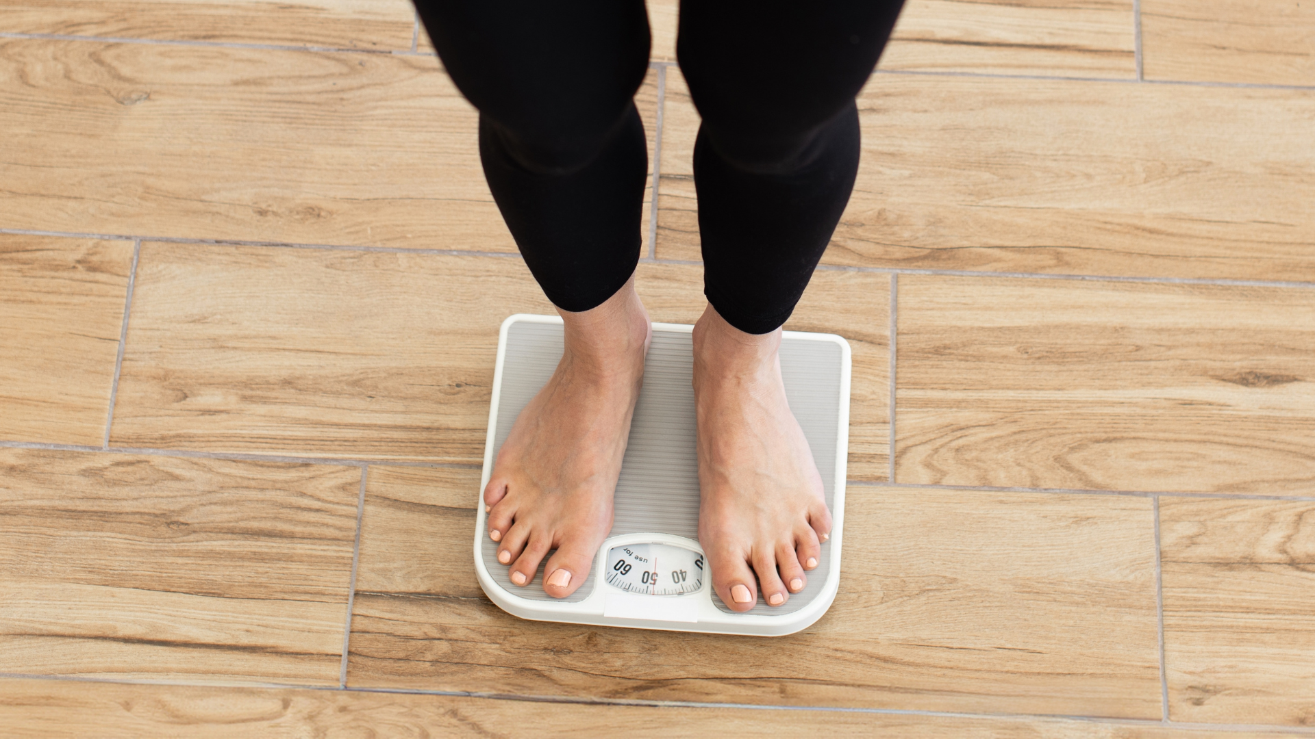 A top-down view of a person standing barefoot on a traditional bathroom scale on a wooden floor.