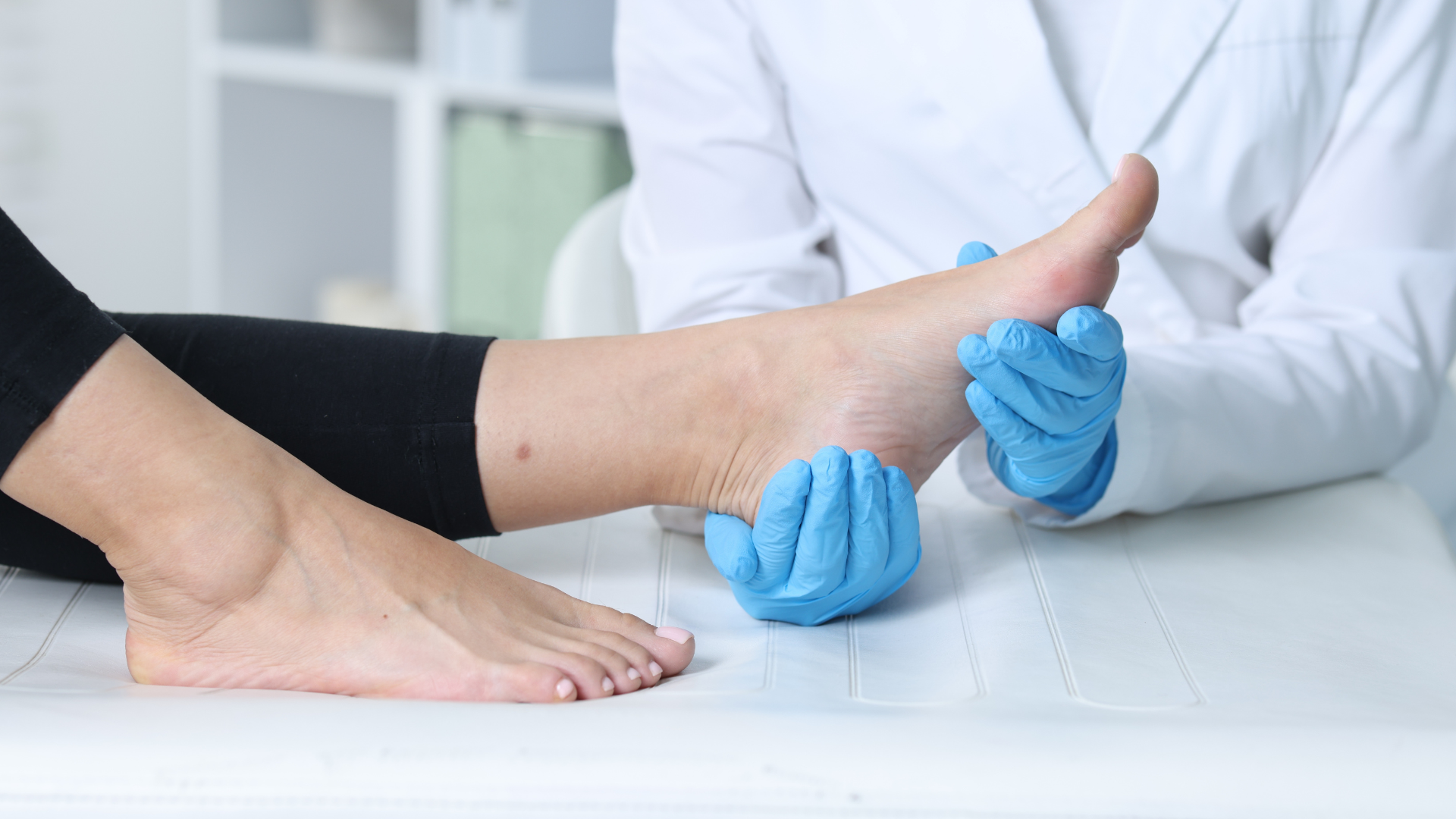A medical professional wearing blue gloves examining the bare foot and ankle of a patient in a clinic.
