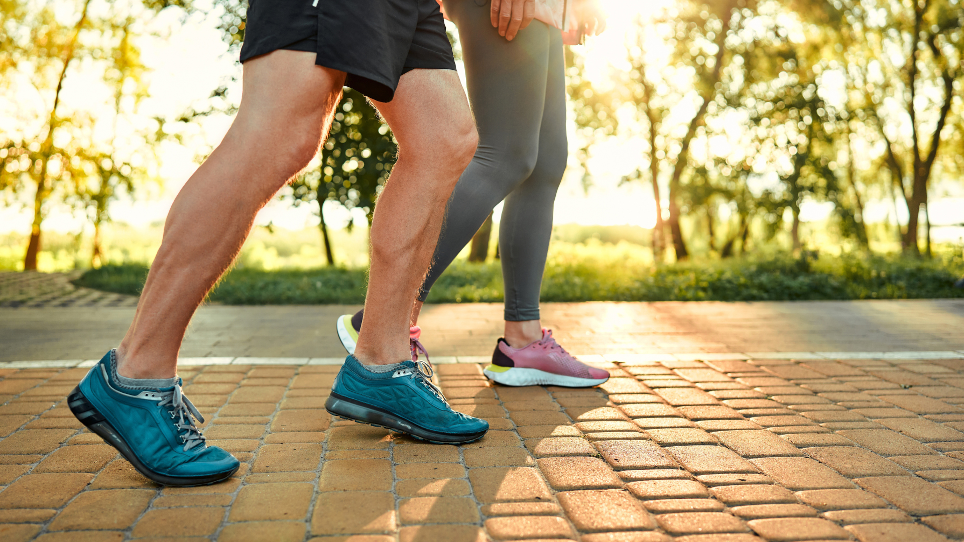 A close-up of the legs of two people wearing athletic clothes and running shoes walking on a paved park path at sunset.