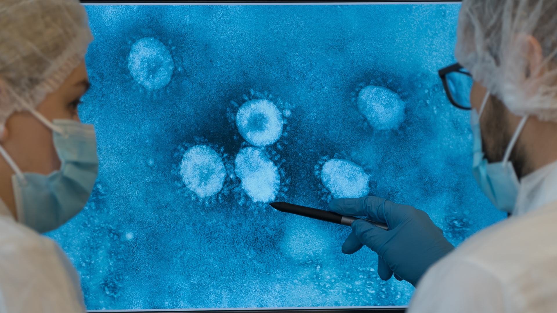Two scientists in medical gear looking at a large blue screen displaying microscopic images of a virus.