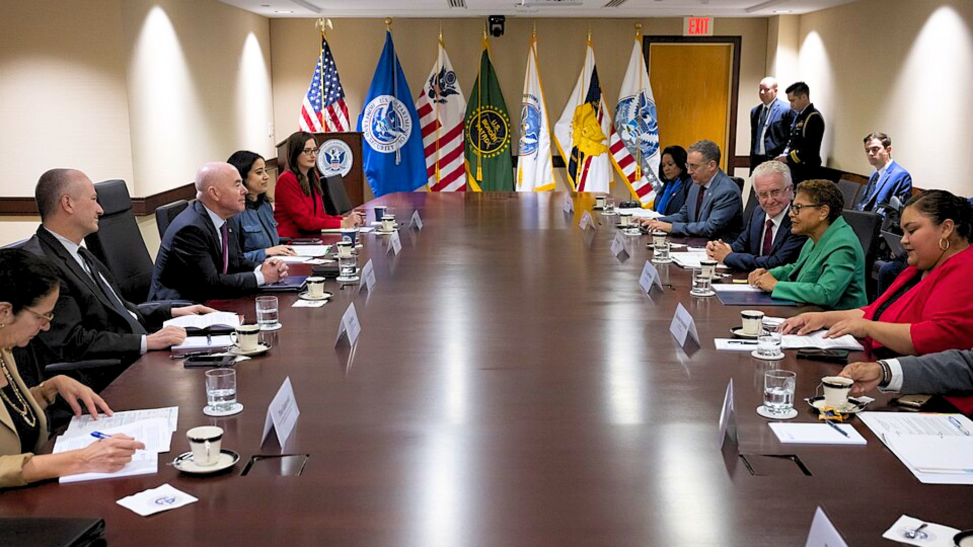 Government officials having a discussion around a long wooden conference table in a meeting room with official flags.