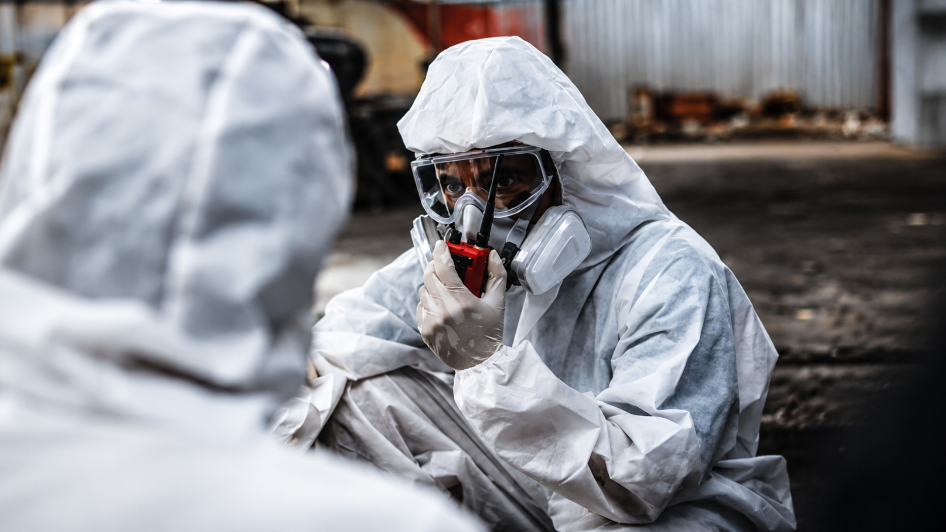 Workers wearing white protective hazmat suits and respirators communicating with a handheld radio during a cleanup.