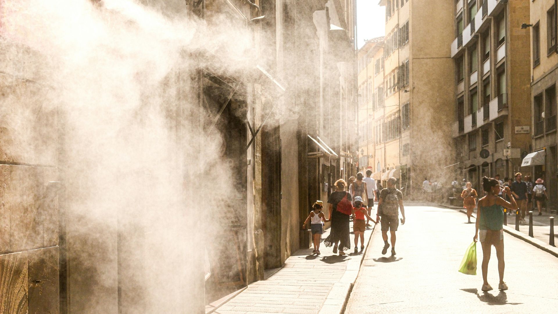 Sunlight streams through a narrow city street as a cooling mist sprays across pedestrians walking between buildings on a hot day.