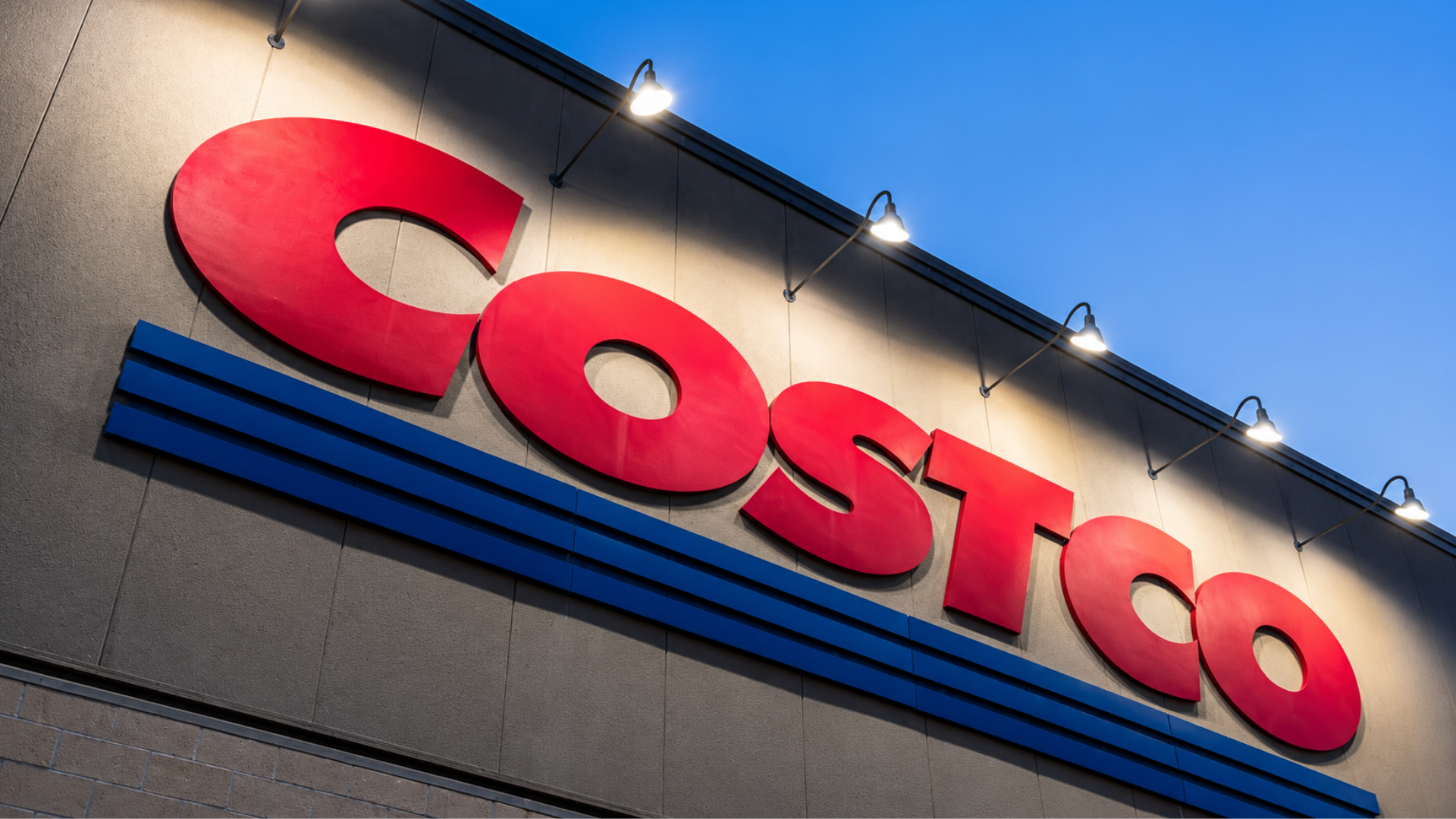 A large, red and blue Costco sign illuminated by spotlights on the exterior wall of a warehouse store.