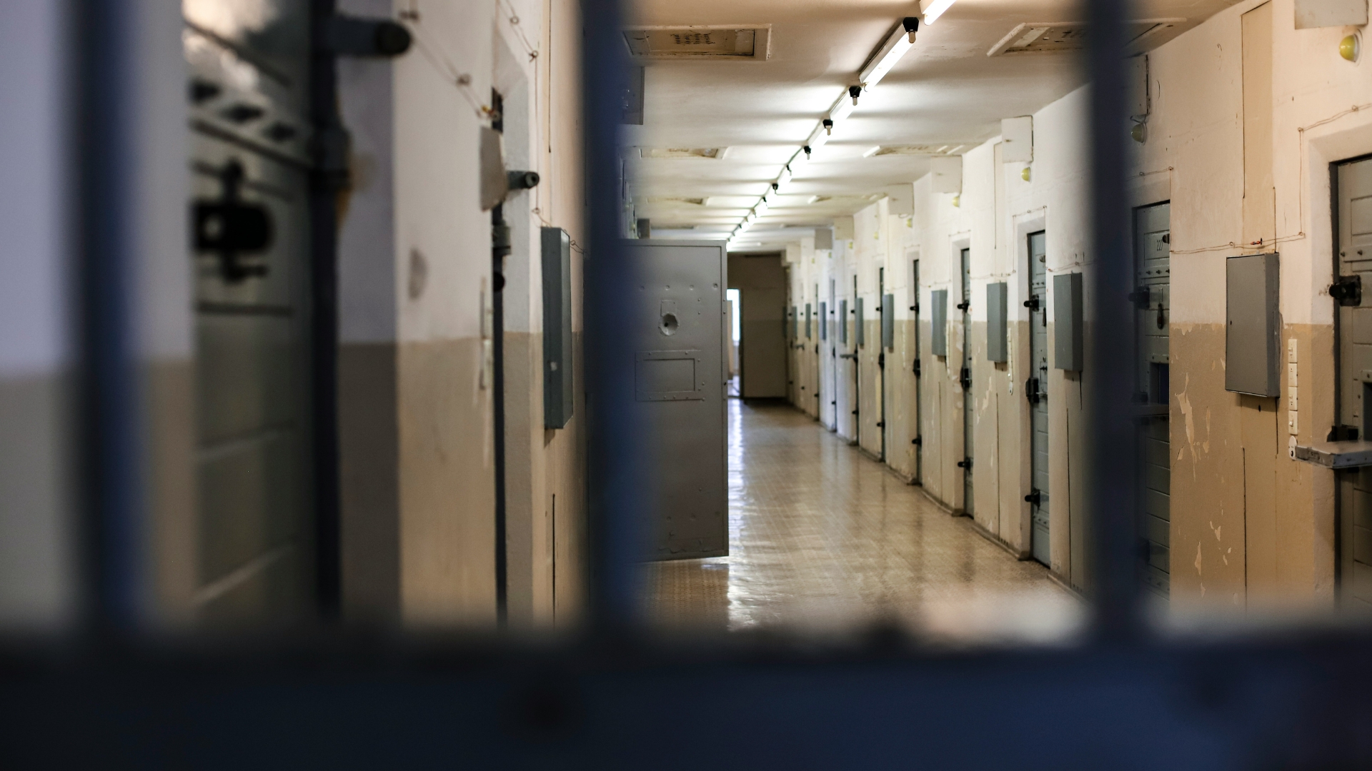 View down a prison cell block through metal bars, showing a long corridor lined with closed cell doors under fluorescent lights.