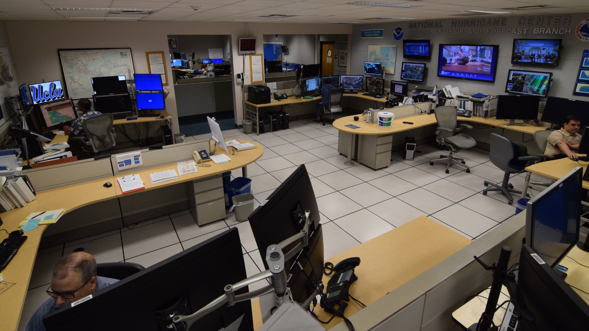 Operations room at the National Hurricane Center with staff working at computer stations surrounded by weather maps and monitoring screens.
