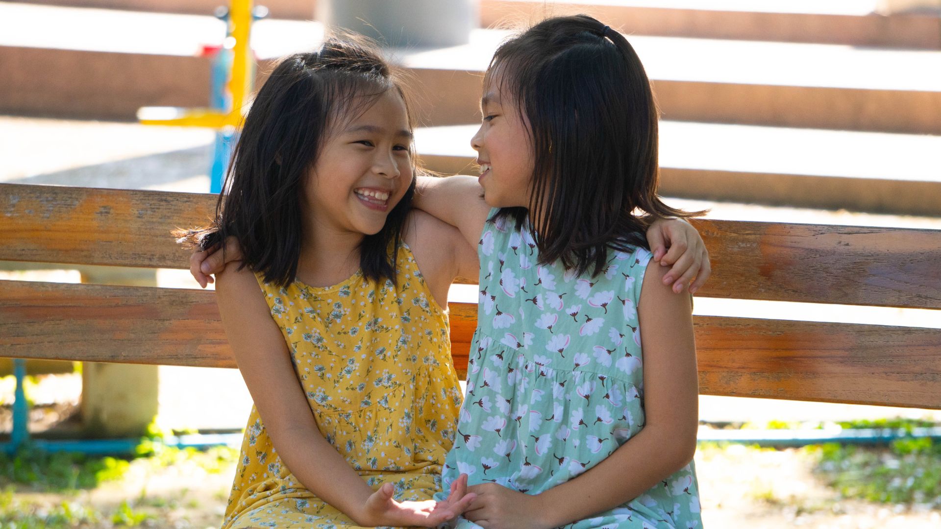 Two little twin sisters sitting on a bench in a street, looking at each other.