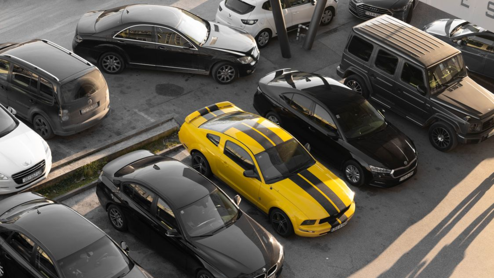 A high-angle shot of an outdoor parking lot where a bright yellow Ford Mustang with dual black racing stripes stands out dramatically among rows of mostly black, grey, and white vehicles.