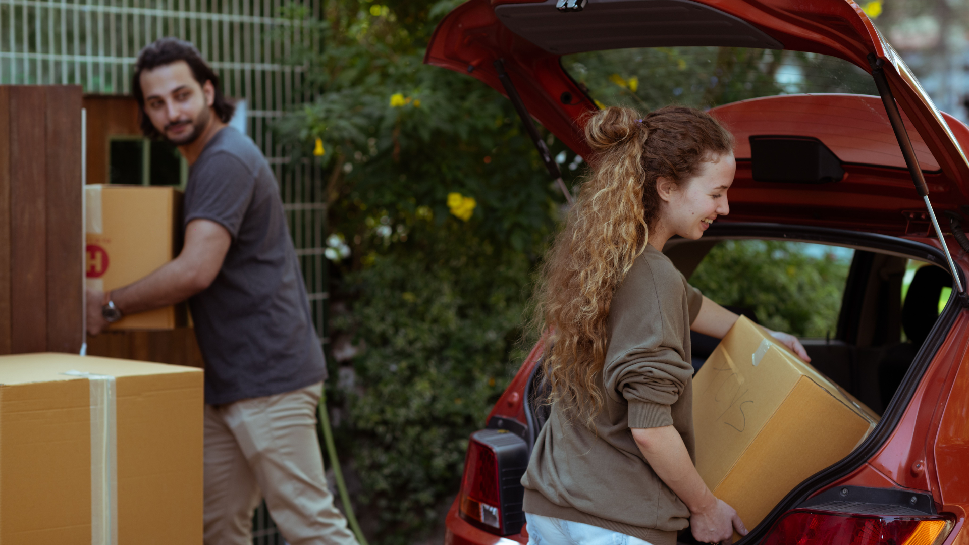 A young couple works together to load brown cardboard moving boxes into the open trunk of a red car parked outside.