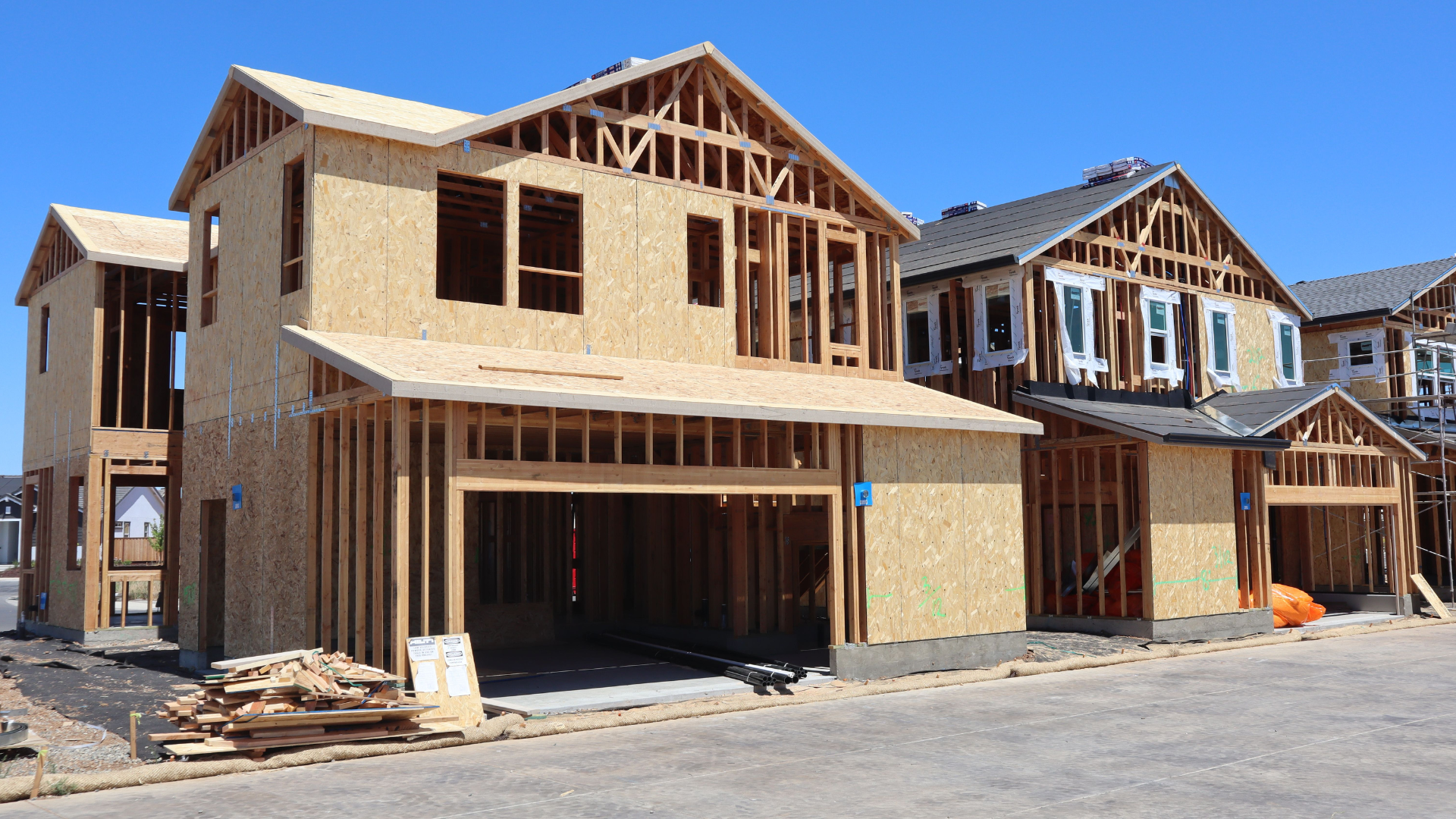 Multiple two-story residential homes stand in a row under a clear blue sky in their wooden framing stage of construction.