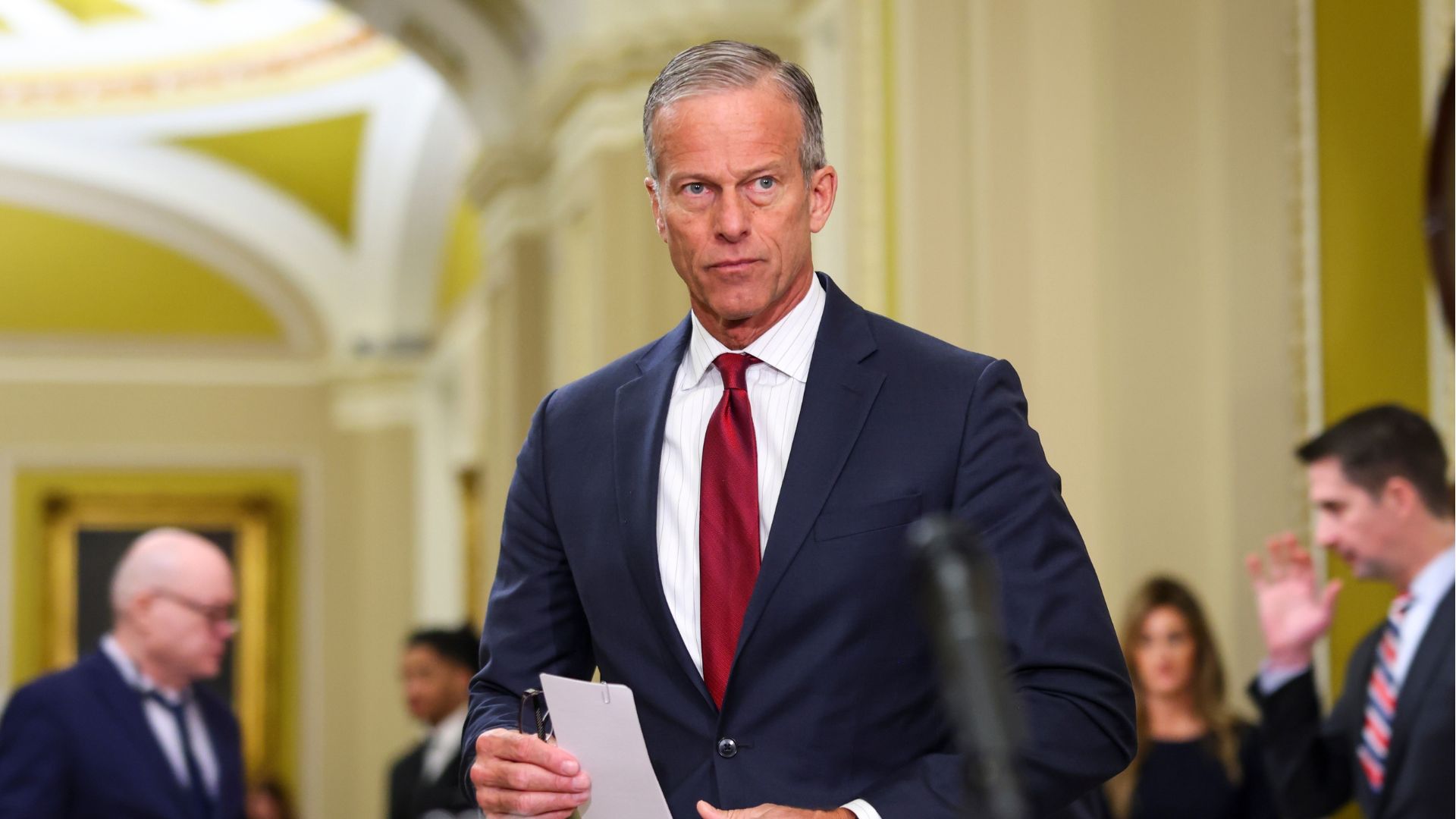 Senate Majority Leader John Thune speaks to reporters, holding a piece of paper and glasses.