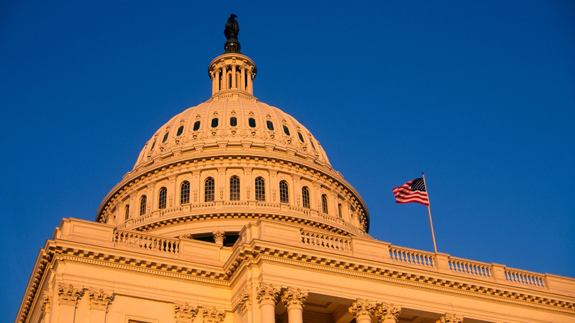 The US Capitol dome under a clear sky with an American flag in the foreground, lit by warm sunlight.