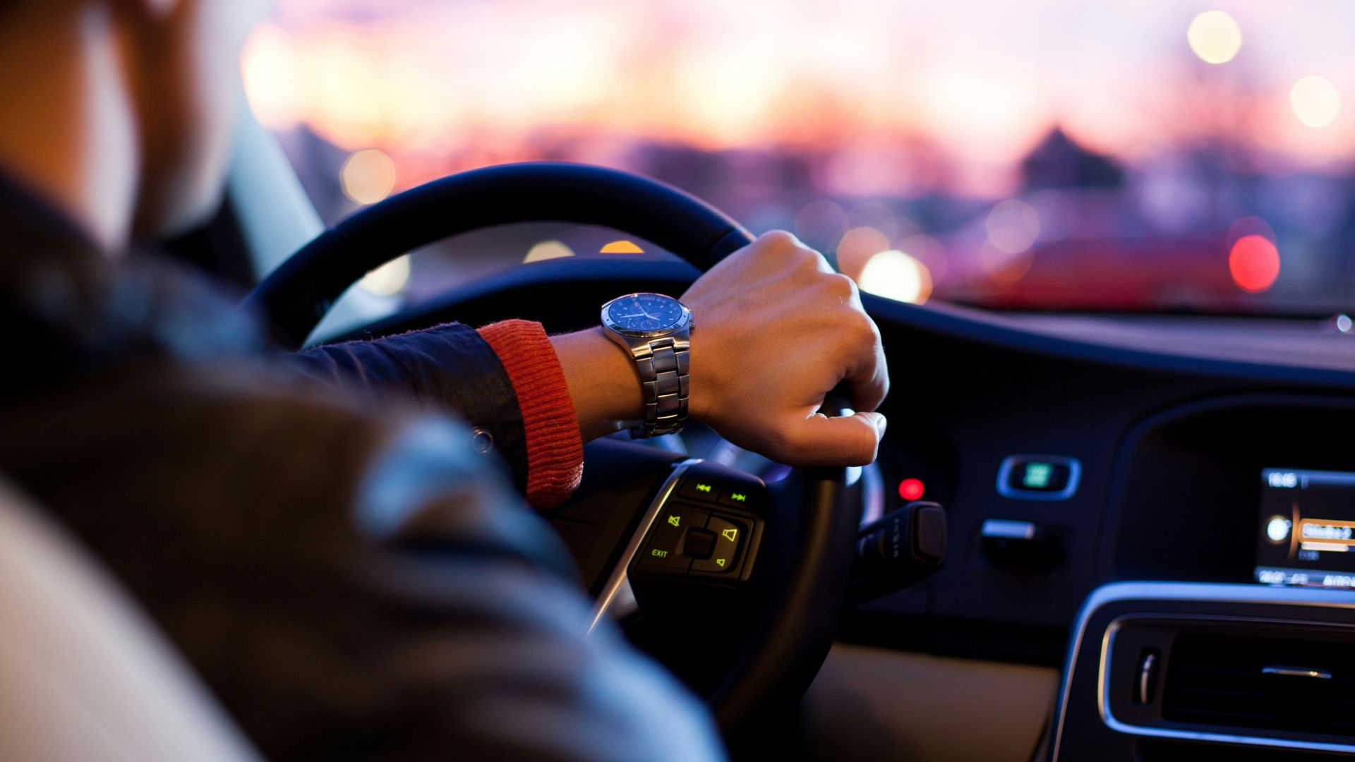 A driver's hand on steering wheel with city skyline at dusk.