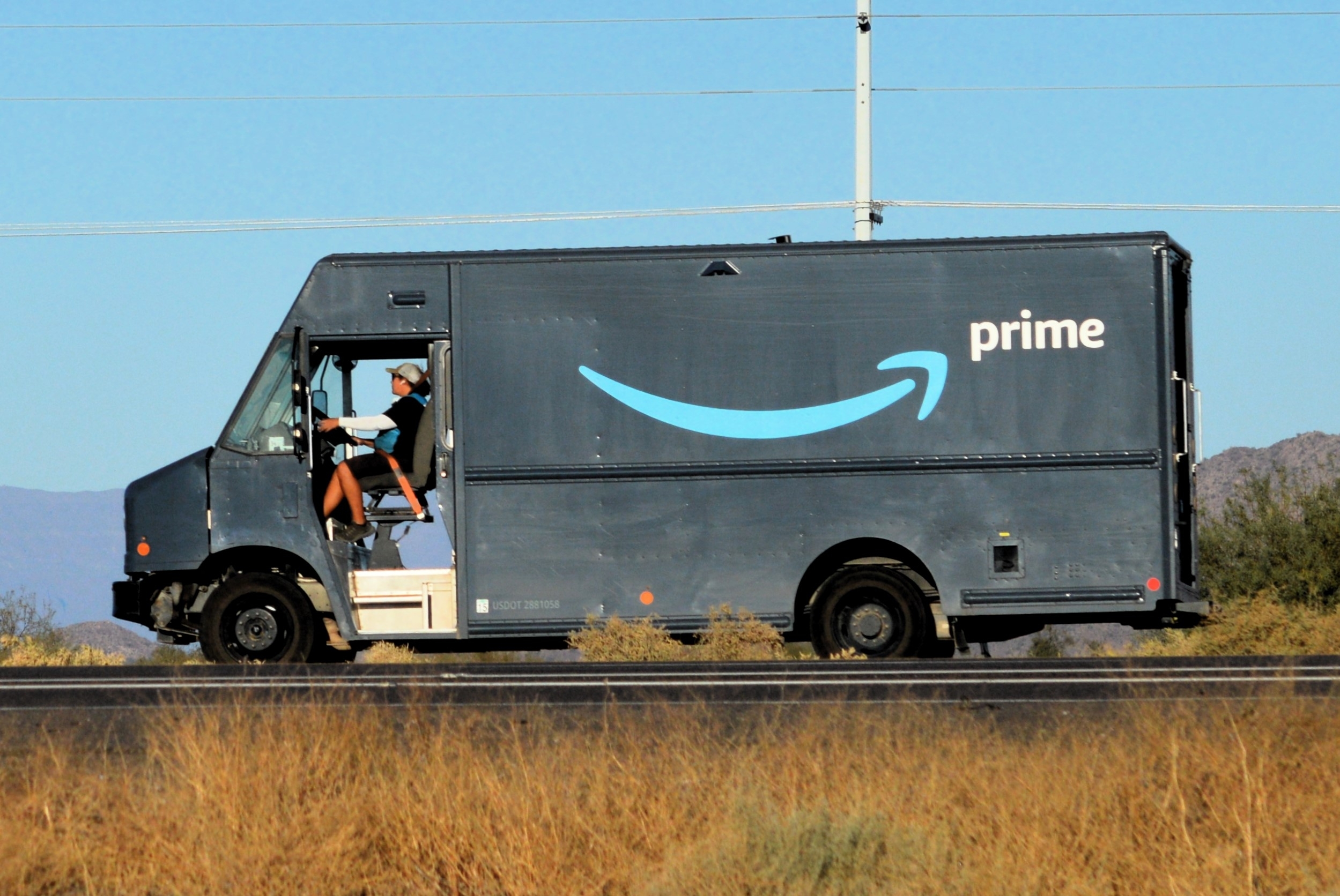 An Amazon driver operates a blue Amazon Prime delivery van traveling along a roadside with dry grass and mountains in the background.