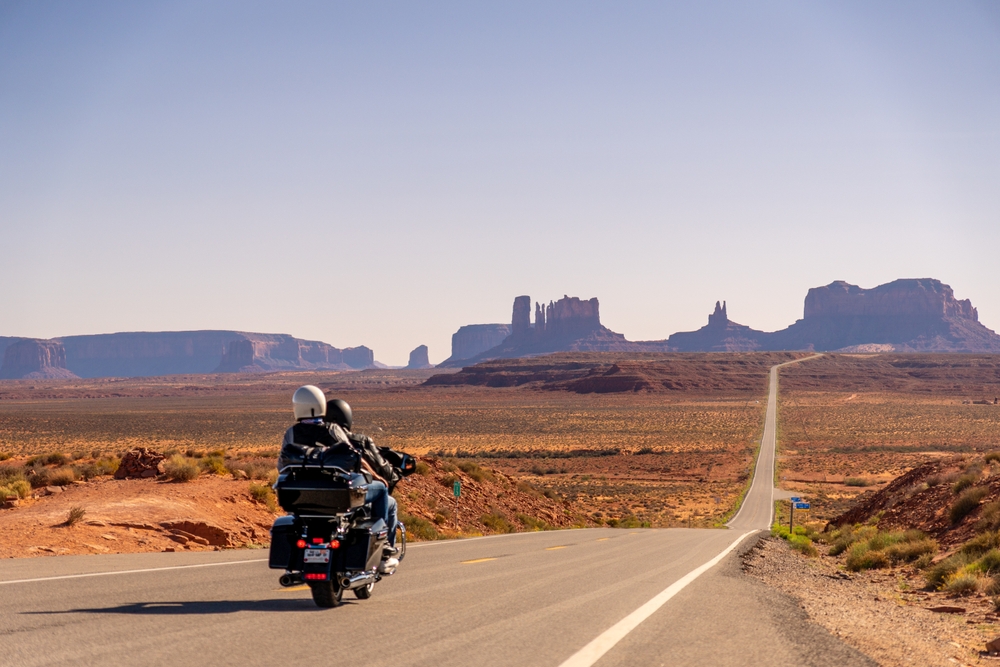 Unrecognizable Couple on Motorcycle at Monument Valley Forest Gump Point down a long, empty road that stretches through the vast, arid desert landscape