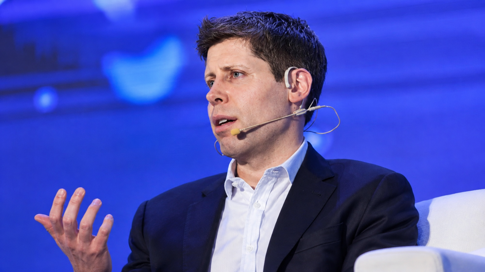 OpenAI CEO Sam Altman speaking on stage at a conference wearing a head-mounted microphone.