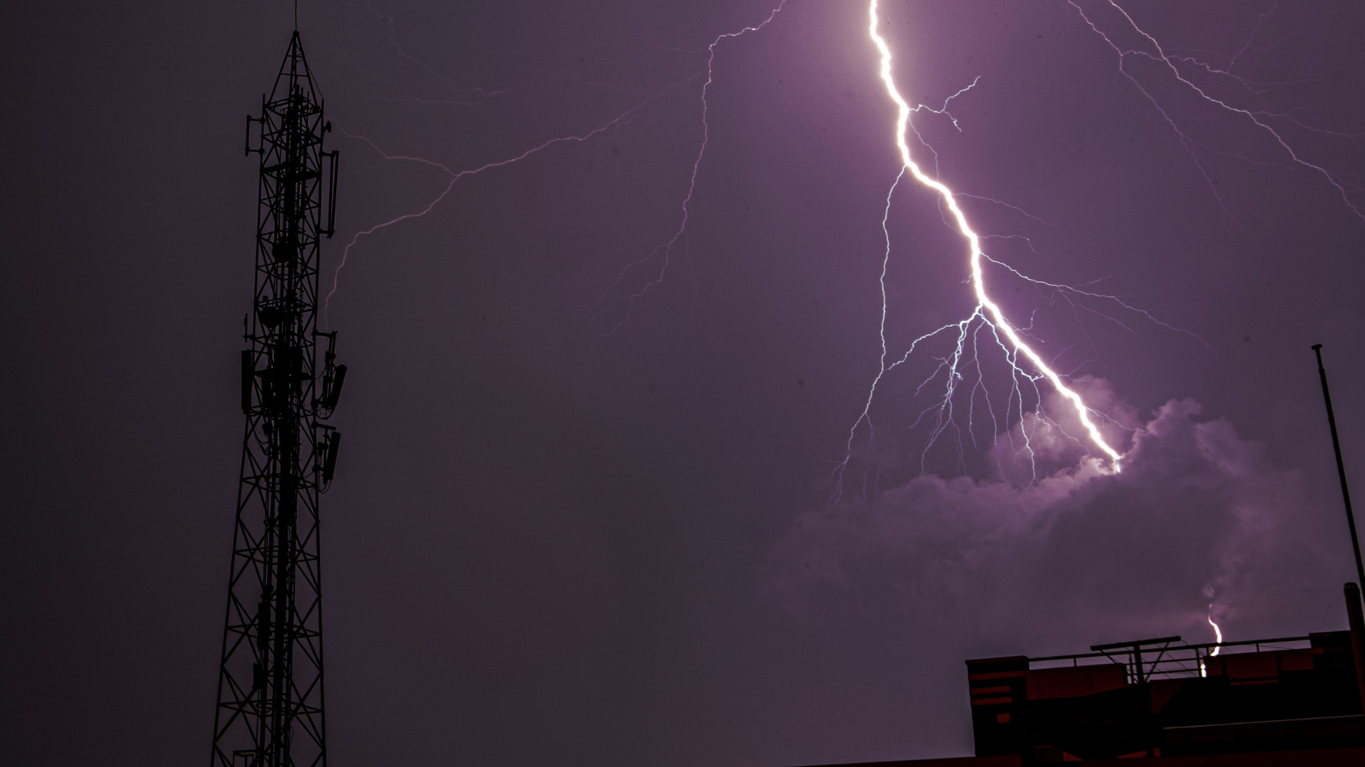 Intense lightning bolt striking near an antenna in a cityscape, captured during a thunderstorm.