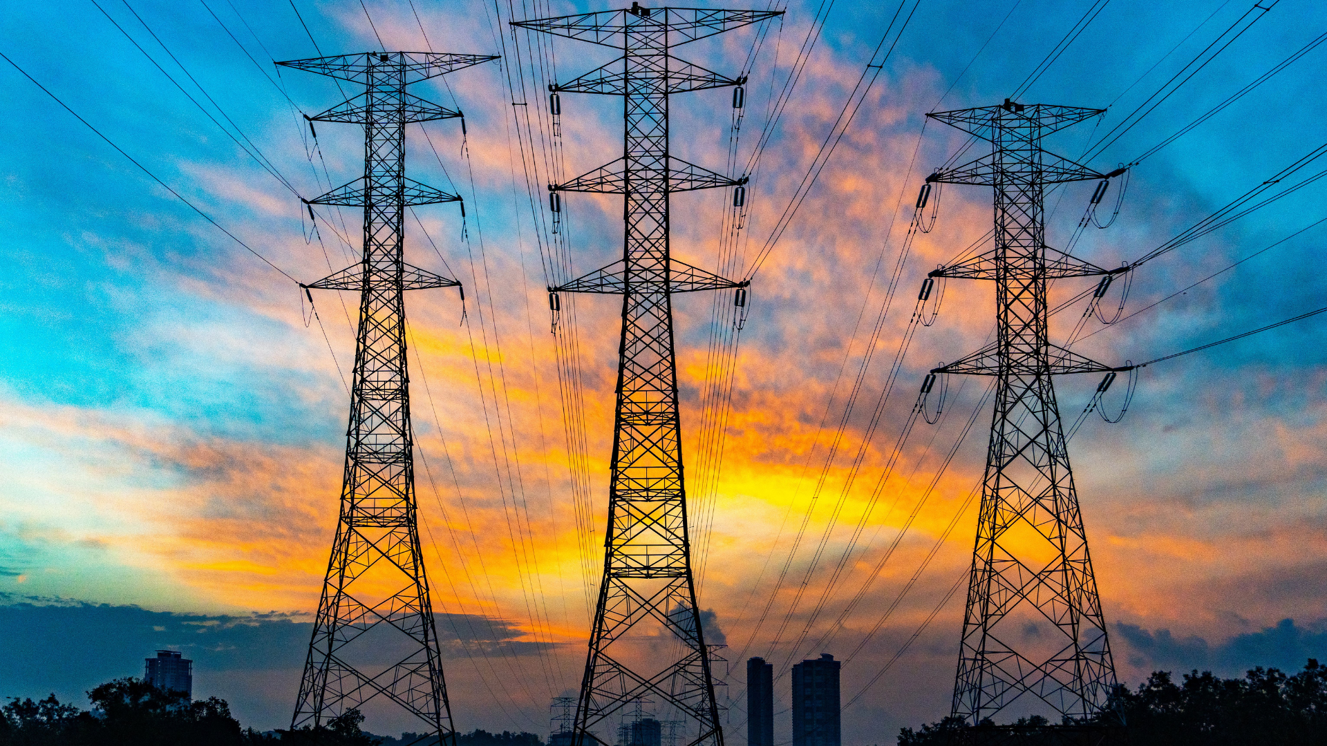 Silhouetted high-voltage transmission towers stretch across a dramatic sunset sky, symbolizing America's strained and underused power grid.