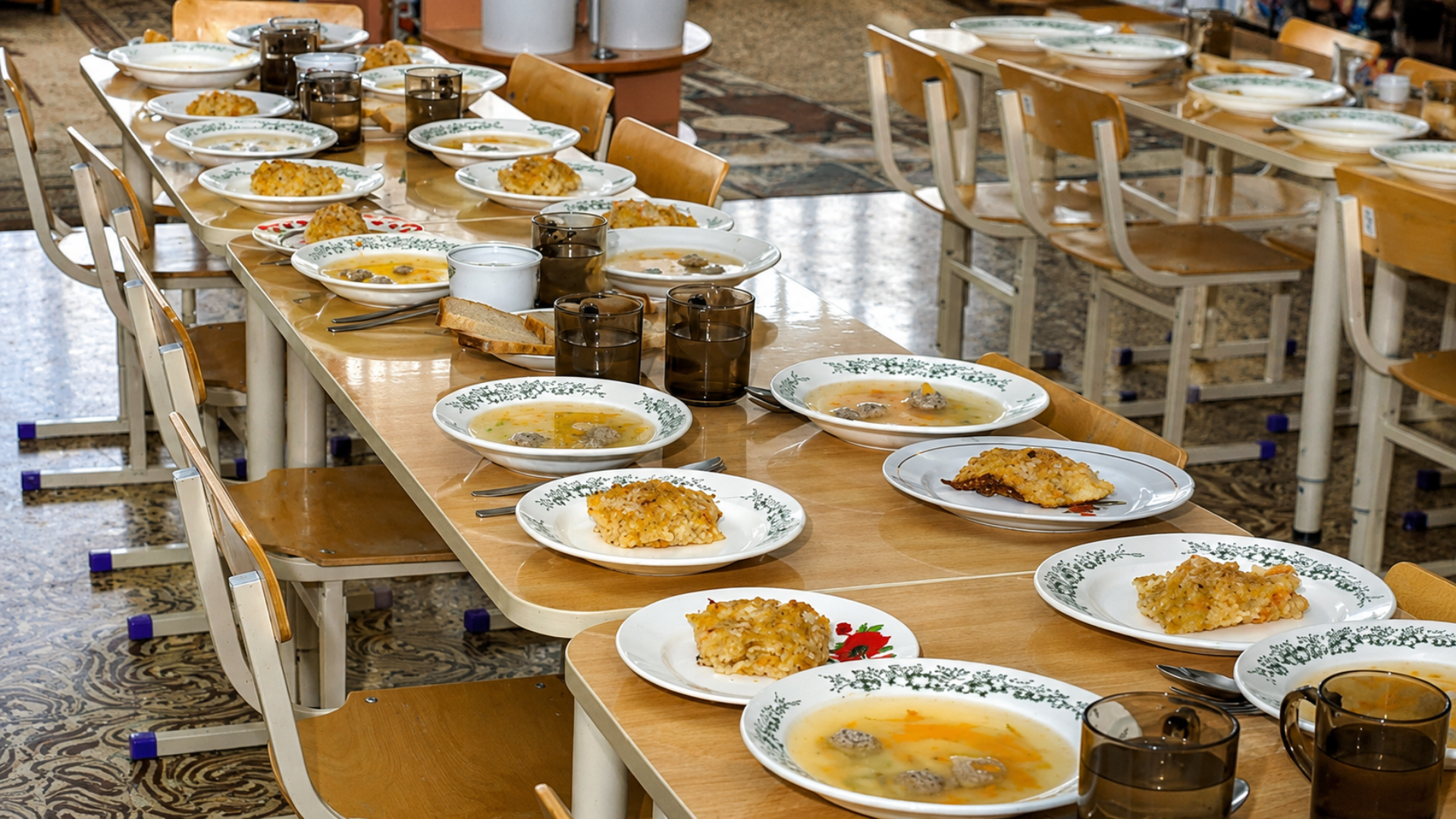 Long cafeteria tables are set with bowls of soup plates of rice casserole bread and dark drinking glasses. Rows of empty wooden chairs surround the prepared meals in a communal dining area.