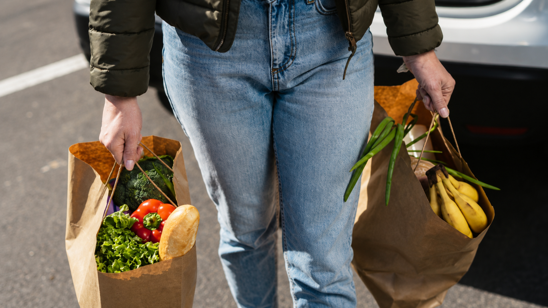 Shopper carrying two paper grocery bags filled with fresh produce and bread.