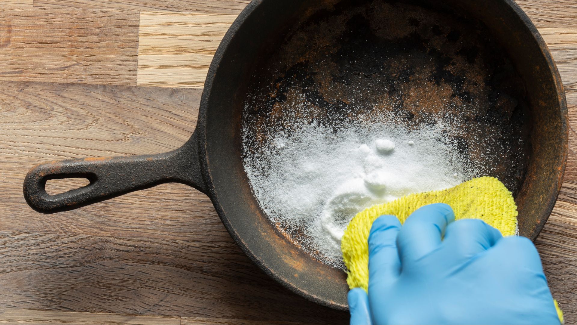 Gloved hand scrubbing a rusty cast-iron skillet with cleaning powder.