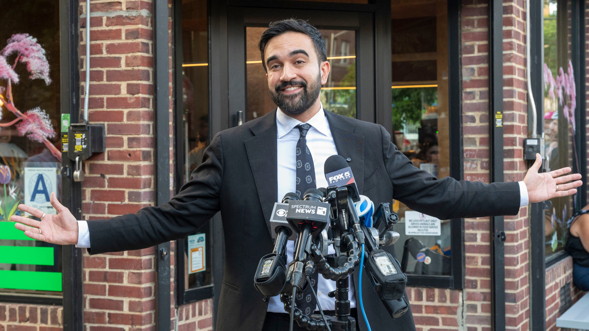 Zohran Mamdani speaking to news microphones outside a brick building.