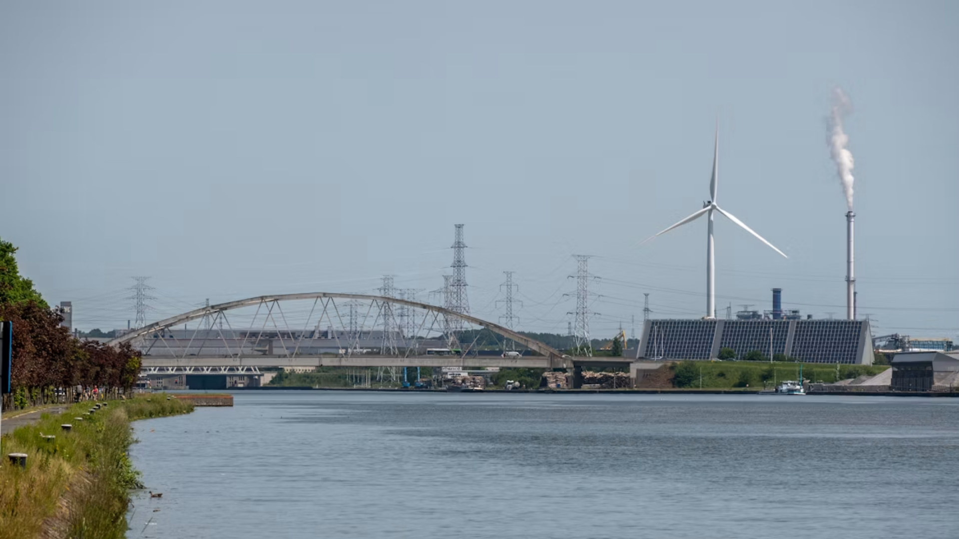 Wind turbine and a gas-fired power plant stand side by side, showing how transmission bottlenecks stall both renewable and fossil fuel energy projects alike.