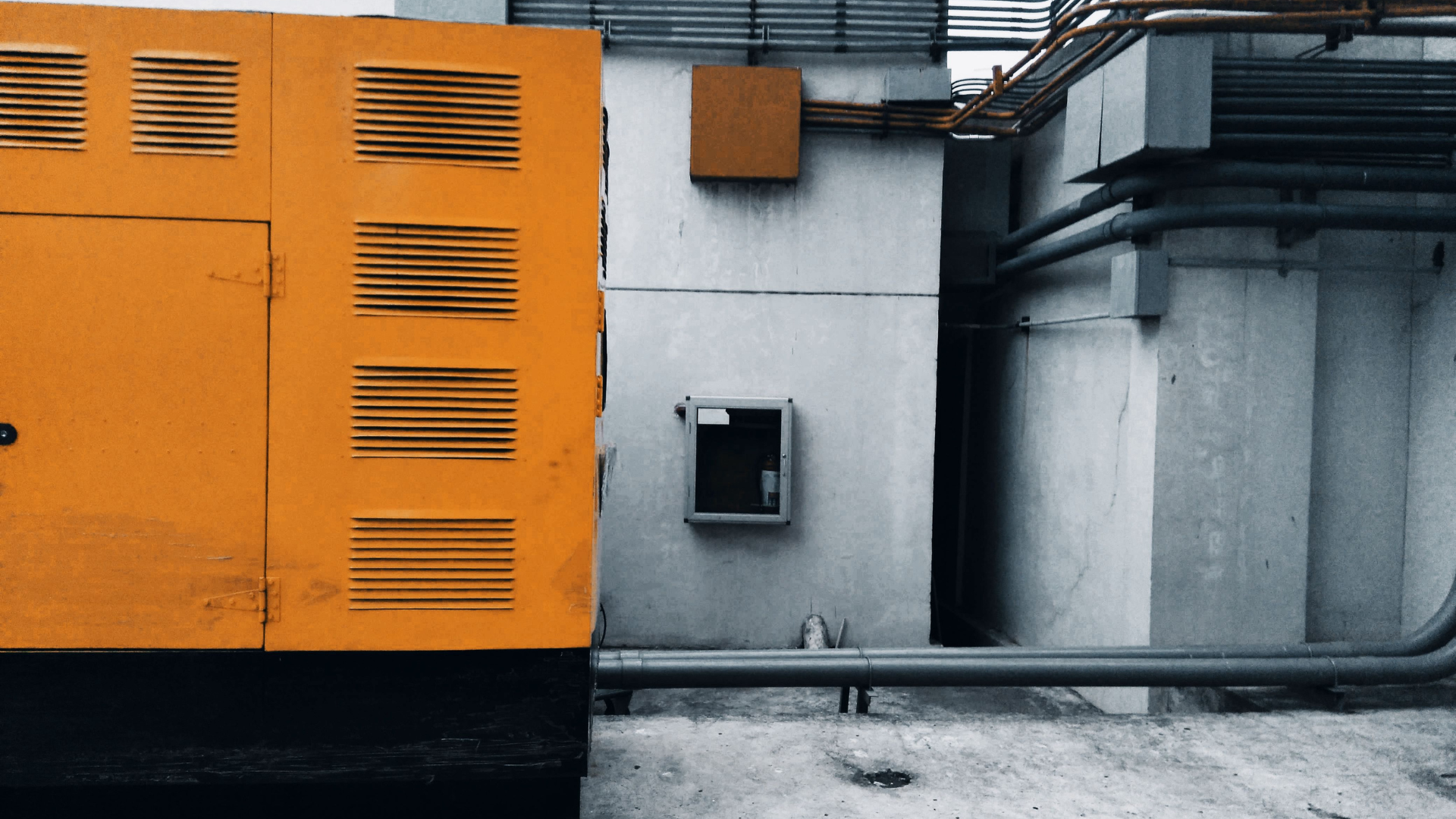 Outdoor view of an industrial generator with visible piping and wiring against a concrete wall.