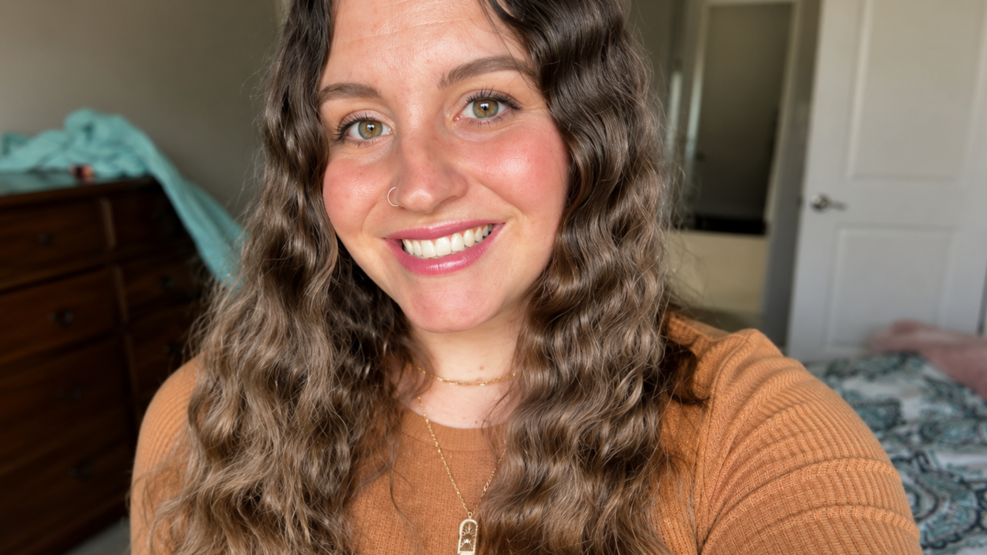 Smiling woman with long wavy brown hair takes a close up selfie in a bedroom. She wears a brown top layered gold necklaces and a small hoop nose ring.
