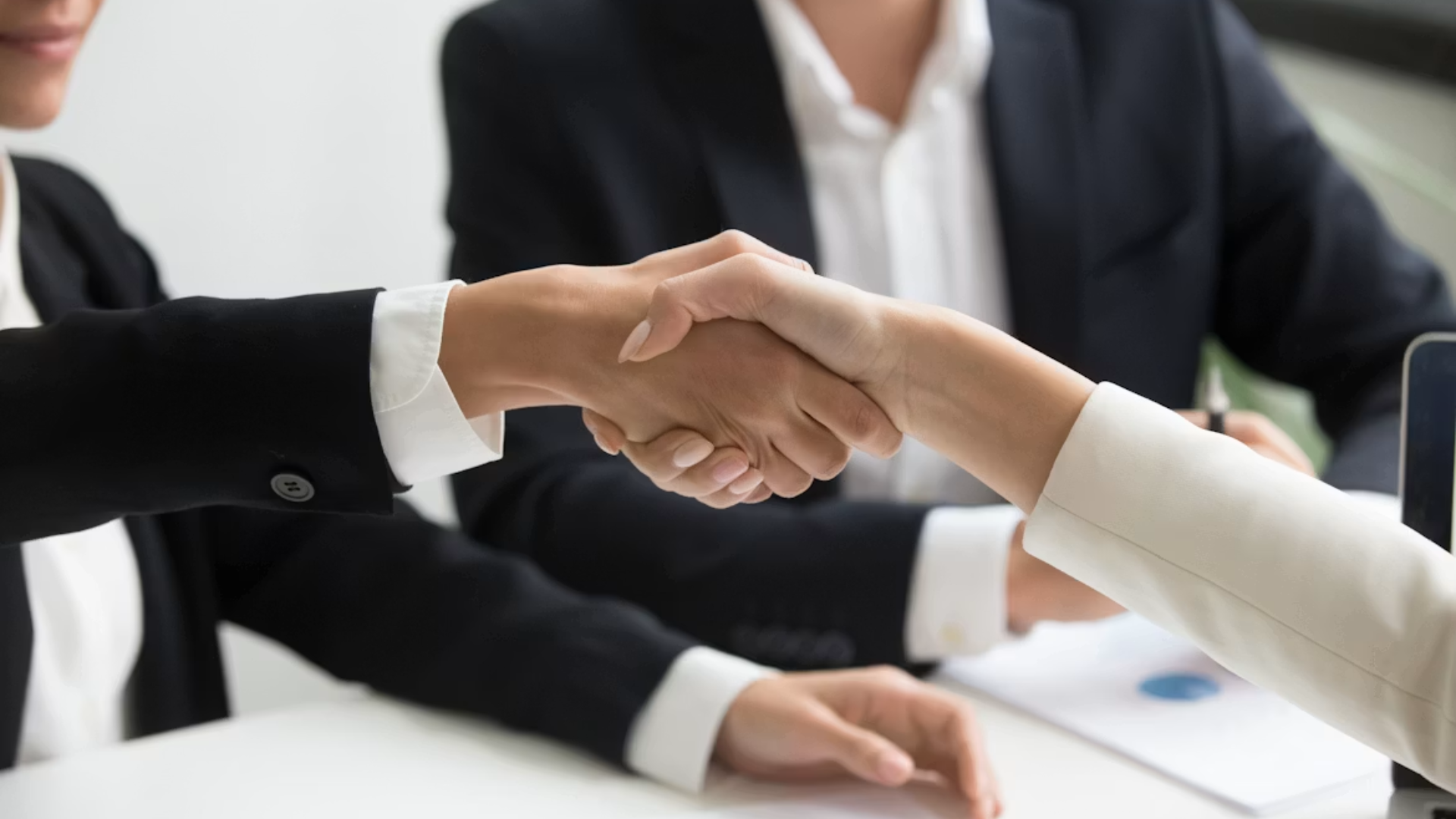 Two business executives shake hands over a signed acquisition document, symbolizing a major corporate ownership change.