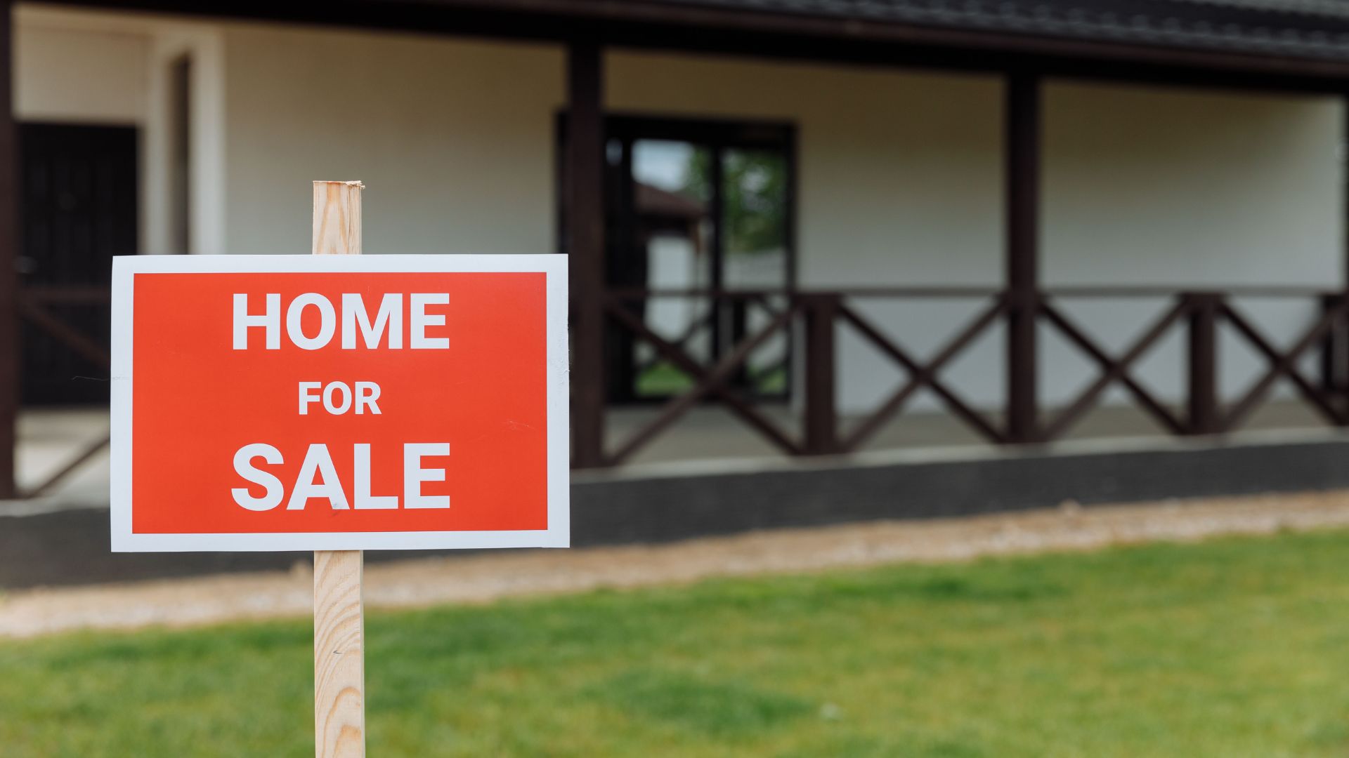 A red 'Home for Sale' sign staked in the front yard of a single-story house with a covered porch.