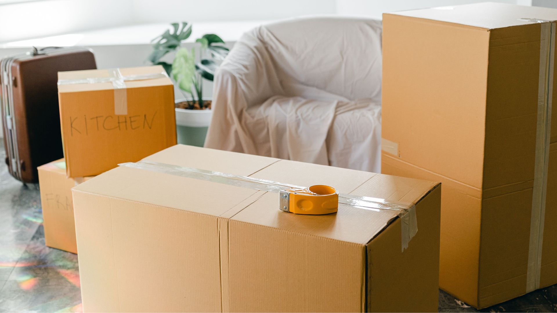 Cardboard moving boxes and a covered chair in a sunlit room.