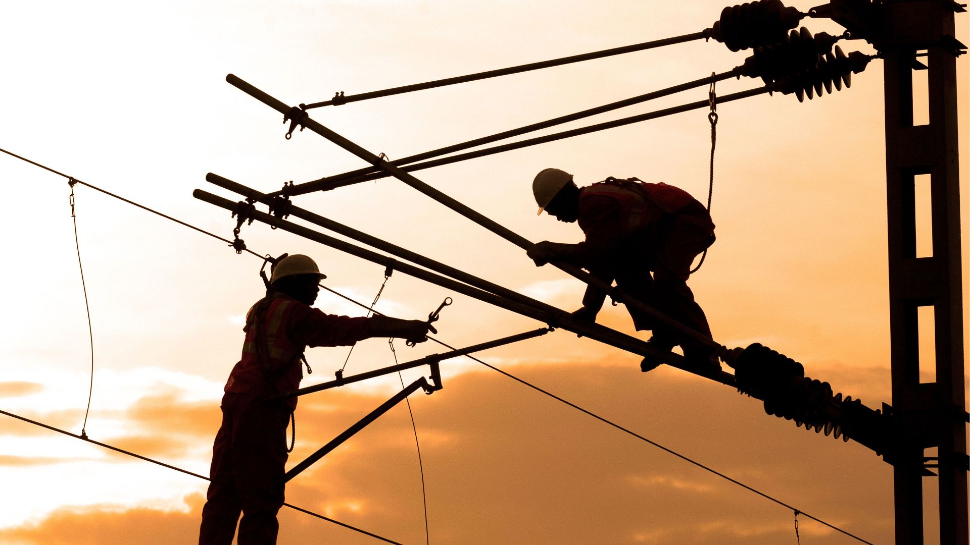 Silhouettes of two utility linemen working on a high-voltage transmission tower against a sunset.