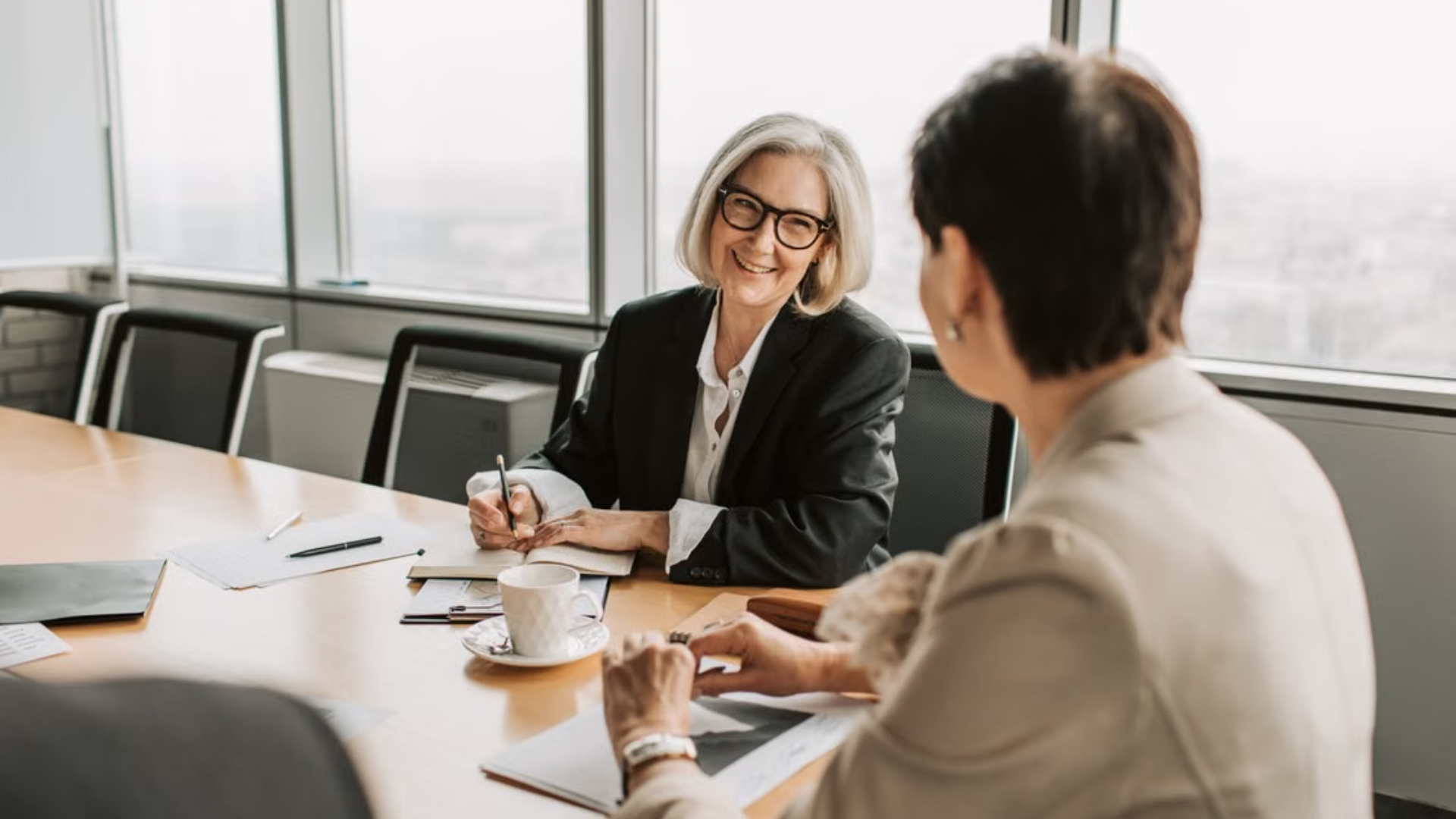  A smiling lawyer with glasses takes notes during a conference room consultation with an older client.
