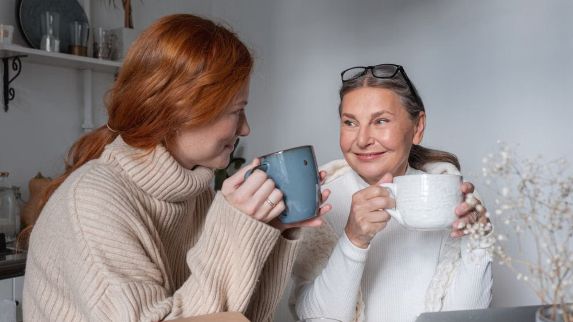 An older woman and a younger woman smile at each other while sharing warm drinks at a table in a home setting.